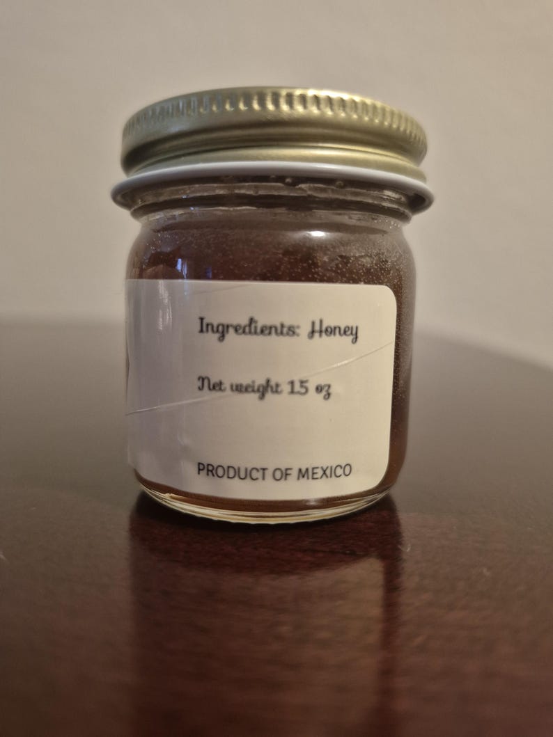 K&ouml;nnte beinhalten: Ein kleines Glas mit goldfarbenem Deckel, gef&uuml;llt mit Honig. Das Etikett lautet "Zutaten: Honig", "Nettogewicht 1,5 oz" und "Produkt aus Mexiko".