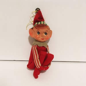 May include: A red felt Christmas elf ornament with a green and white striped hat, brown painted face, and white and red striped arms.