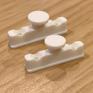 May include: Two white plastic door latch catches with a round knob. The catches have two holes on each side.