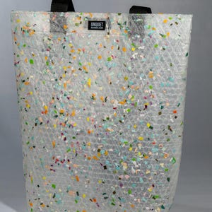 May include: A clear tote bag with a bubble wrap texture and colourful confetti-like accents. The bag has black handles and a small label that reads "UNQUIET SUSTAINABLE DESIGN". The bag is rectangular and stands upright.
