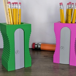 May include: Two pencil sharpeners, one green and one pink, with a gray sharpening blade. Each sharpener has a compartment for pencils. The sharpeners are on a wooden surface.