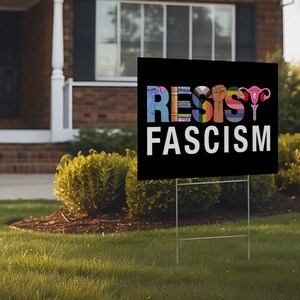 May include: Two yellow and black yard signs with the text "Resist Fascism" in colorful letters. The letters are made up of various symbols, including a fist, a uterus, and a rainbow.