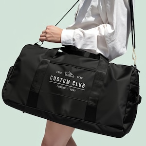 May include: A black duffel bag with a shoulder strap featuring a white graphic print that reads ESTD. YEAR CUSTOM CLUB CUSTOM TEXT with a small sneaker icon. The bag has black nylon webbing handles and a gold-toned metal clip on the strap.
