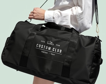 May include: A black duffel bag with a shoulder strap featuring a white graphic print that reads ESTD. YEAR CUSTOM CLUB CUSTOM TEXT with a small sneaker icon. The bag has black nylon webbing handles and a gold-toned metal clip on the strap.