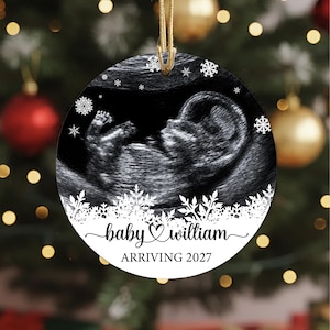 May include: A round, flat ornament featuring a black and white ultrasound image of a fetus. The design includes white snowflake graphics, the text "baby William" in a cursive script with a heart, and the printed text "ARRIVING 2027" below.