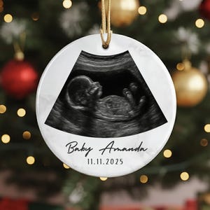 May include: A white, round Christmas ornament with a gold string. The ornament features a black and white ultrasound image of a baby. The text "Baby Amanda" and the date "11.11.2025" are printed on the ornament.