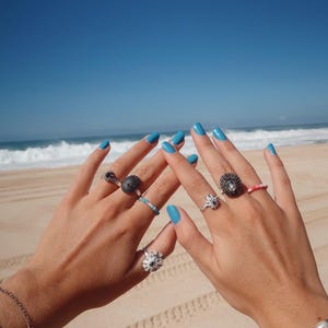 May include: Two hands with blue painted nails, adorned with various rings. The rings feature unique designs, including floral and textured patterns. A beach and ocean backdrop provides a bright, sunny setting for the jewelry.