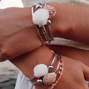 May include: A collection of silver and gold bracelets featuring white and brown seashells and colourful beads. The bracelets are stacked, creating a layered effect. The seashells give the jewellery a beachy, coastal feel.
