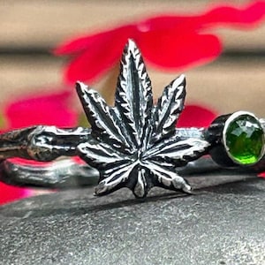 May include: A silver ring featuring a detailed marijuana leaf design and a green gemstone. The ring is displayed against a dark grey background.