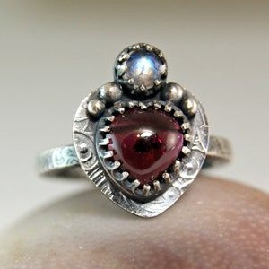May include: A silver ring with a heart-shaped red gemstone and a smaller, round, iridescent gemstone. The ring is decorated with intricate details.