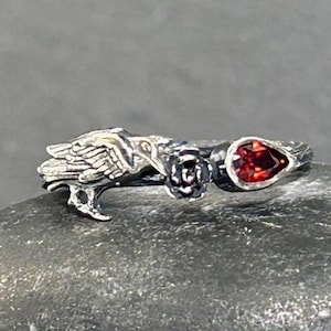 May include: A silver ring featuring a bird, a rose, and a teardrop-shaped red gemstone. The ring has intricate details and a nature-inspired design. The gemstone is approximately 0.3 inches in length.