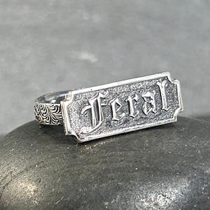 May include: A silver-toned ring featuring a rectangular plaque with the word Feral embossed in a gothic-style font. The plaque has a textured, dark background, and the ring band is decorated with an intricate, repeating floral scrollwork pattern.