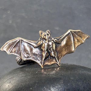 May include: A metallic ring featuring a detailed, sculpted bat with outstretched wings. The bat has a dark, antiqued finish that highlights the texture of its fur and wing structure. The ring band is visible beneath the bat, showing intricate patterns.