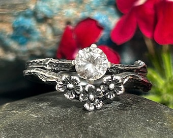 May include: A silver ring set with a large, clear, round gemstone. The band is made to look like a branch with small flower accents.