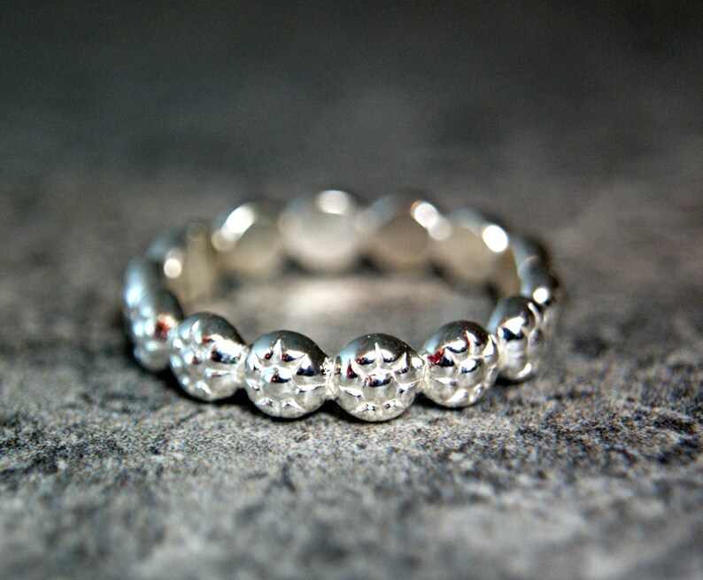 May include: A silver ring with a textured, beaded design. The ring is made of a smooth, polished metal and has a simple, elegant style.