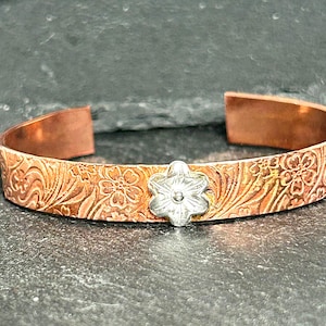 May include: Copper cuff bracelet with a floral pattern and a silver-colored flower accent. The bracelet has an open design and is displayed on a dark surface. The cuff is approximately 0.75 inches wide.