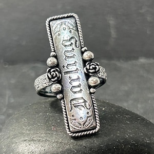 Puede incluir: Un anillo plateado con la palabra "Darling" grabada verticalmente. El anillo presenta un centro rectangular con detalles ornamentados, enmarcado por un diseño de cuerda trenzada. Pequeños acentos de rosas y cuentas adornan los lados, agregando un toque de elegancia.