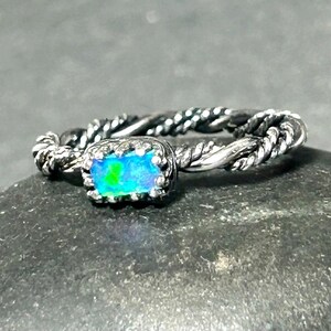 May include: A silver ring with a twisted band design. The ring features a rectangular, iridescent gemstone with blue and green hues. The gemstone is set in a decorative bezel.