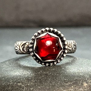 Puede incluir: Un anillo de plata con una piedra preciosa roja engastada en un engaste de bisel. El anillo tiene una banda decorativa con diseños florales.