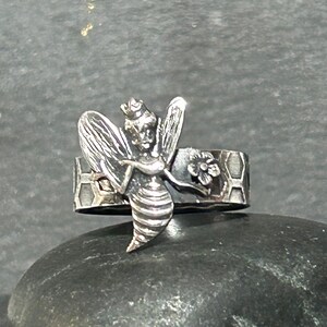 May include: A silver-toned ring featuring a detailed bee design. The bee has outstretched wings and a crown. The ring band has a honeycomb pattern and a small flower. The ring is set against a dark background.