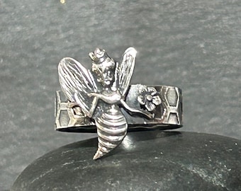 May include: A silver-toned ring featuring a detailed bee design. The bee has outstretched wings and a crown. The ring band has a honeycomb pattern and a small flower. The ring is set against a dark background.