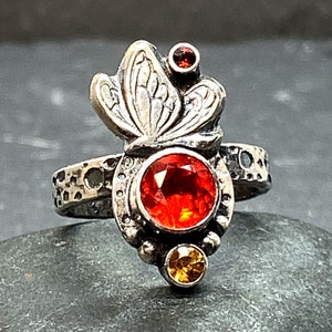 May include: A silver ring with a butterfly design and three gemstones. The largest gemstone is a red orange, the middle gemstone is a yellow, and the smallest gemstone is a red.