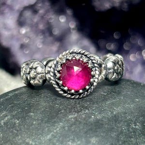Könnte beinhalten: Ein silberner Ring mit einem rosafarbenen Edelstein, der in einer dekorativen Fassung sitzt. Der Ring hat eine strukturierte Band mit floralen Designs.