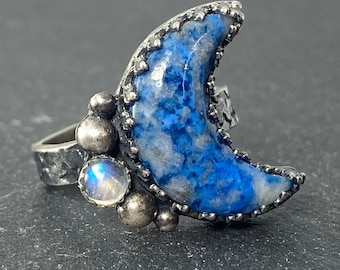 May include: A silver ring featuring a crescent moon shaped blue and white lapis lazuli stone. The ring is adorned with small silver beads and a moonstone.