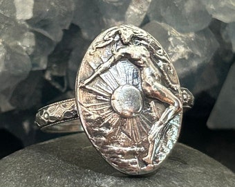 Könnte beinhalten: Ein silberner Ring mit einem ovalen Design, das eine Frauenfigur zeigt, die auf einer Sonne mit Strahlen steht. Der Ring hat ein detailliertes, strukturiertes Design.