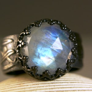 May include: A silver ring with a large, round, faceted moonstone. The moonstone has blue and white internal reflections. The ring has a dark, ornate bezel and a detailed floral design on the band.