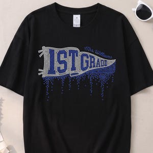 May include: A black short-sleeved t-shirt featuring a rhinestone-embellished pennant design that reads 1ST GRADE and Mrs. Williams in blue and clear crystals, with a dripping effect at the bottom of the graphic and a white tag at the neckline.
