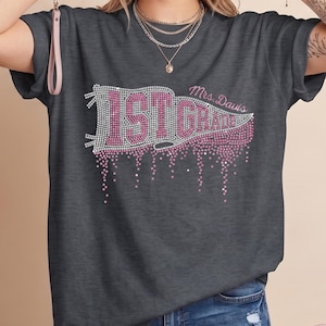 May include: A charcoal gray short-sleeve t-shirt featuring a rhinestone graphic of a pennant flag with the text 1ST GRADE and Mrs. Davis. The design includes a dripping effect created with pink and clear rhinestones across the front chest area.
