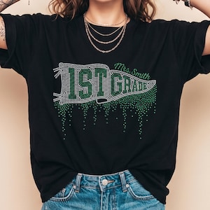 Pode incluir: Uma t-shirt preta de manga curta com um gráfico de galhardete adornado com strass que diz 1ST GRADE em verde e branco. O design inclui o texto Mrs. Smith e um efeito de gotejamento criado com strass verdes na parte inferior.