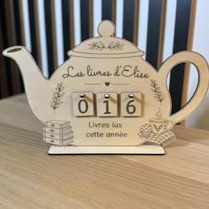 Può includere: Un cartello in legno chiaro a forma di teiera con il testo Les livres d'Elise e Livres lus cette année. Include tre blocchi quadrati rotanti che mostrano i numeri 0, 1 e 6, insieme a illustrazioni incise di una pila di libri, un muffin e una tazza da tè.