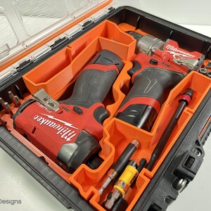 May include: A black and orange tool case containing two red and black power drills and various drill bits. The drills are labelled "Milwaukee". The case has a clear lid and orange interior compartments.