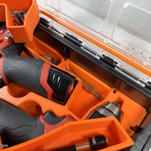 May include: An orange plastic tool case with a clear lid. The case contains two red and black cordless drills, a drill bit, and a screwdriver bit. The drills have the word "Milwaukee" printed on them.