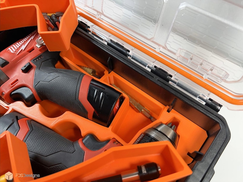May include: An orange plastic tool case with a clear lid. The case contains two red and black cordless drills, a drill bit, and a screwdriver bit. The drills have the word "Milwaukee" printed on them.
