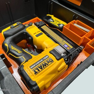 Peut inclure: Un cloueur sans fil DeWalt 20V sans balais jaune et noir avec une poign&eacute;e noire et une batterie, &agrave; l'int&eacute;rieur d'un organisateur d'outils orange et noir. L'organisateur comporte plusieurs compartiments de rangement.