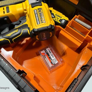 Peut inclure: Un cloueur sans fil DeWalt 20V jaune et noir et un bloc-batterie sont dans un organisateur d'outils orange et noir. Un paquet d'agrafes de calibre 23 de 2,54 cm est &eacute;galement visible.