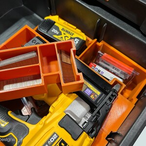 Peut inclure: Une bo&icirc;te &agrave; outils noire ouverte avec des compartiments de rangement orange. &Agrave; l'int&eacute;rieur se trouvent un cloueur jaune et noir, une batterie jaune et diverses fixations. Les compartiments contiennent des agrafes et de petits morceaux de bois.