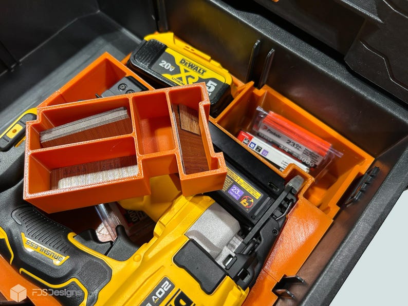 Peut inclure: Une bo&icirc;te &agrave; outils noire ouverte avec des compartiments de rangement orange. &Agrave; l'int&eacute;rieur se trouvent un cloueur jaune et noir, une batterie jaune et diverses fixations. Les compartiments contiennent des agrafes et de petits morceaux de bois.