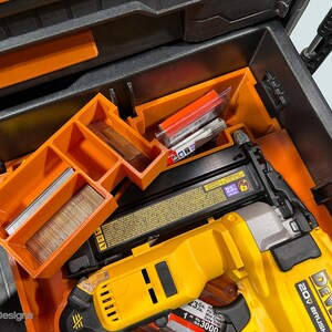 Peut inclure: Une bo&icirc;te &agrave; outils noire ouverte avec des compartiments orange et un cloueur Dewalt 20V Brushless jaune et noir. La bo&icirc;te contient divers clous et fixations, rang&eacute;s dans des plateaux orange.