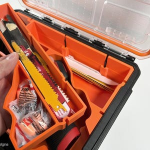 May include: An orange and black toolbox with a clear lid open. The toolbox is filled with various tools, including a saw blade, earplugs, and a small knife. The toolbox is made of durable plastic and has a sturdy handle.