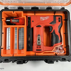 May include: An open orange and black carrying case with a red Milwaukee brand electric staple gun, a black and orange battery, and a compartment with rows of silver staples.