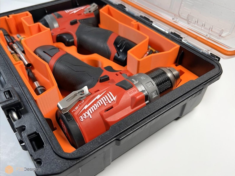 May include: Two red and black cordless drills with the Milwaukee logo are stored in a black and orange hard case with a clear lid.