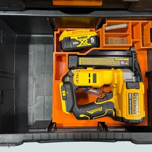 Peut inclure: Une mallette &agrave; outils noire et orange ouverte, contenant un cloueur jaune et noir, une batterie et des compartiments de rangement. Le cloueur porte l'inscription "DEWALT 20V BRUSHLESS".