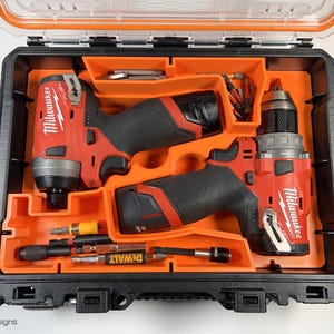 May include: Two Milwaukee power drills, one red and black impact driver and one red and black drill, are stored in a black and orange hard plastic case. The case has a compartment for storing additional drill bits and accessories. The case is open and the drills are visible.