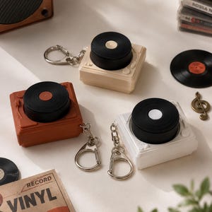 Könnte beinhalten: Drei quadratische Etuis, die wie Miniatur-Plattenspieler gestaltet sind und jeweils eine schwarze, kreisförmige Vinylplatte auf der Oberseite aufweisen. Die Etuis sind in Beige, Terrakotta und Weiß erhältlich und haben einen silbernen Schlüsselanhänger.