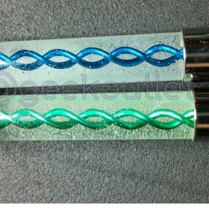 Puede incluir: Dos tubos de vidrio con tapas plateadas. Los tubos contienen un l&iacute;quido transparente con una espiral azul y verde en su interior.