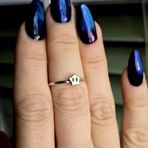 Könnte beinhalten: Ein silberner Ring mit einem sternförmigen Anhänger. Der Stern ist weiß mit goldenen Konturen. Der Ring befindet sich an einem Finger mit dunkelblauem Nagellack.